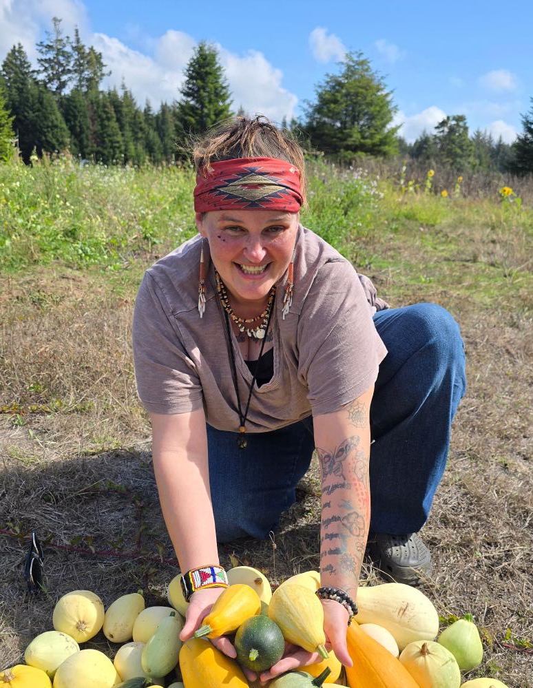 A seedkeeper kneeling with an armful of squash and vegetables from the harvest